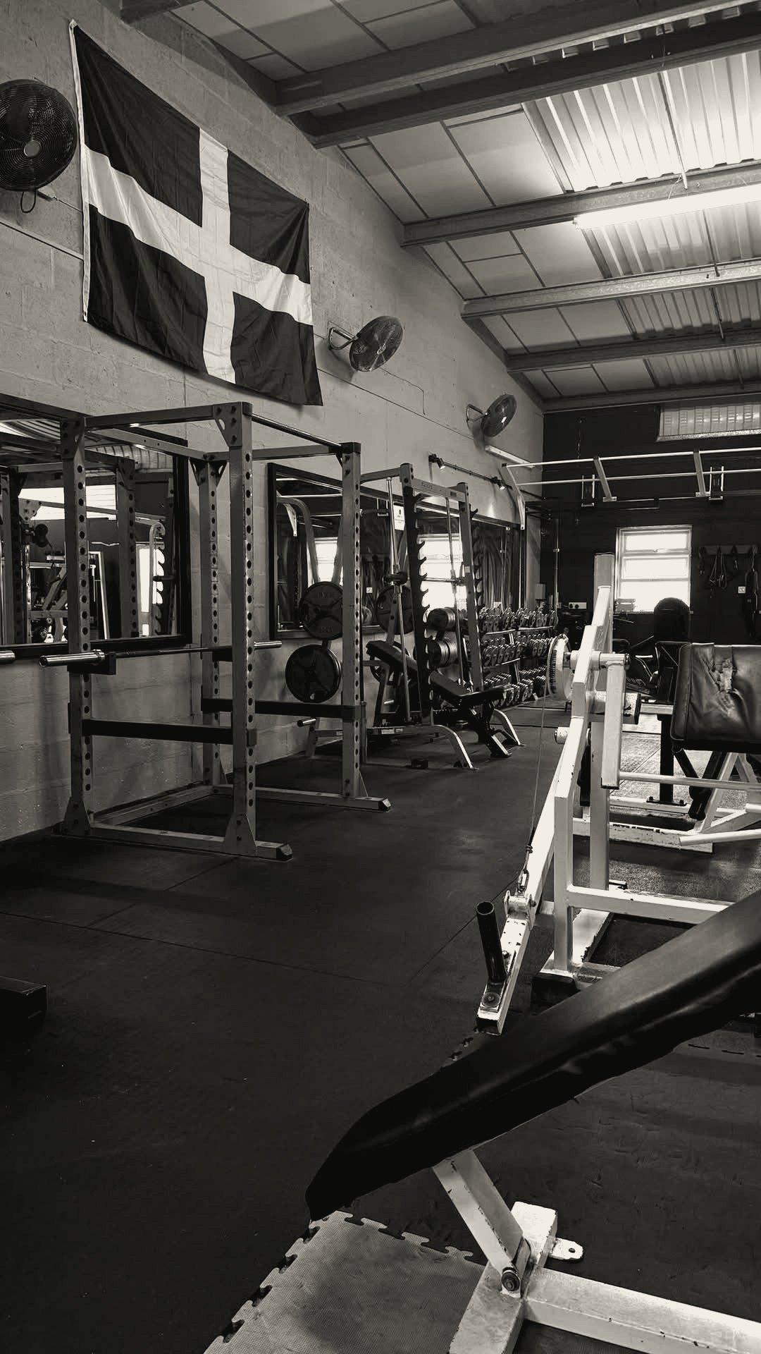 The gym floor with power racks and a plate-loaded machine, and the St Piran flag on the wall